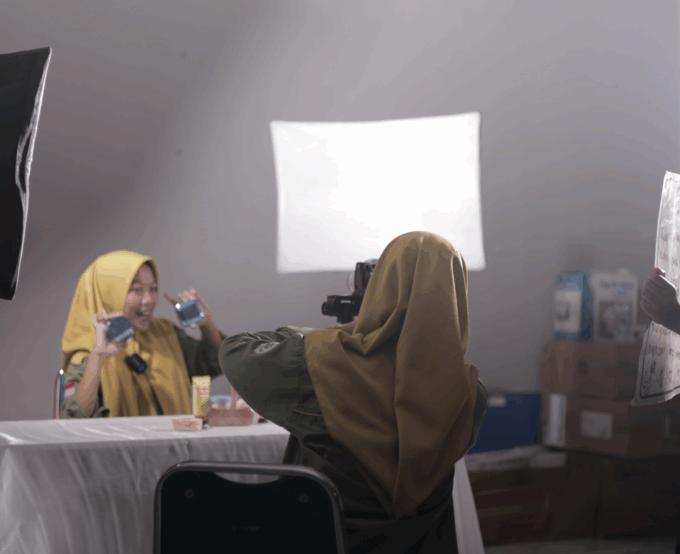 Siswa SMK Muhammadiyah 2 Bantul Program Keahlian Pemasaran sedang praktik live selling di kelas studio. Seorang siswa berperan sebagai host mempresentasikan produk skincare di depan kamera, sementara siswa lain mengoperasikan kamera dan pencahayaan profesional untuk mendukung siaran langsung di media sosial.