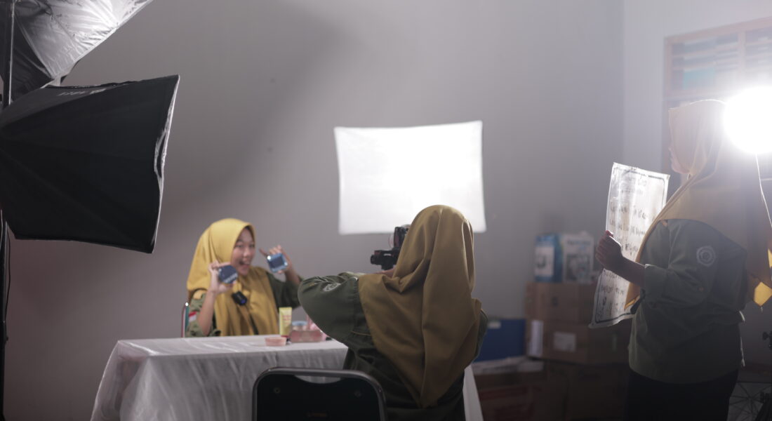 Siswa SMK Muhammadiyah 2 Bantul Program Keahlian Pemasaran sedang praktik live selling di kelas studio. Seorang siswa berperan sebagai host mempresentasikan produk skincare di depan kamera, sementara siswa lain mengoperasikan kamera dan pencahayaan profesional untuk mendukung siaran langsung di media sosial.